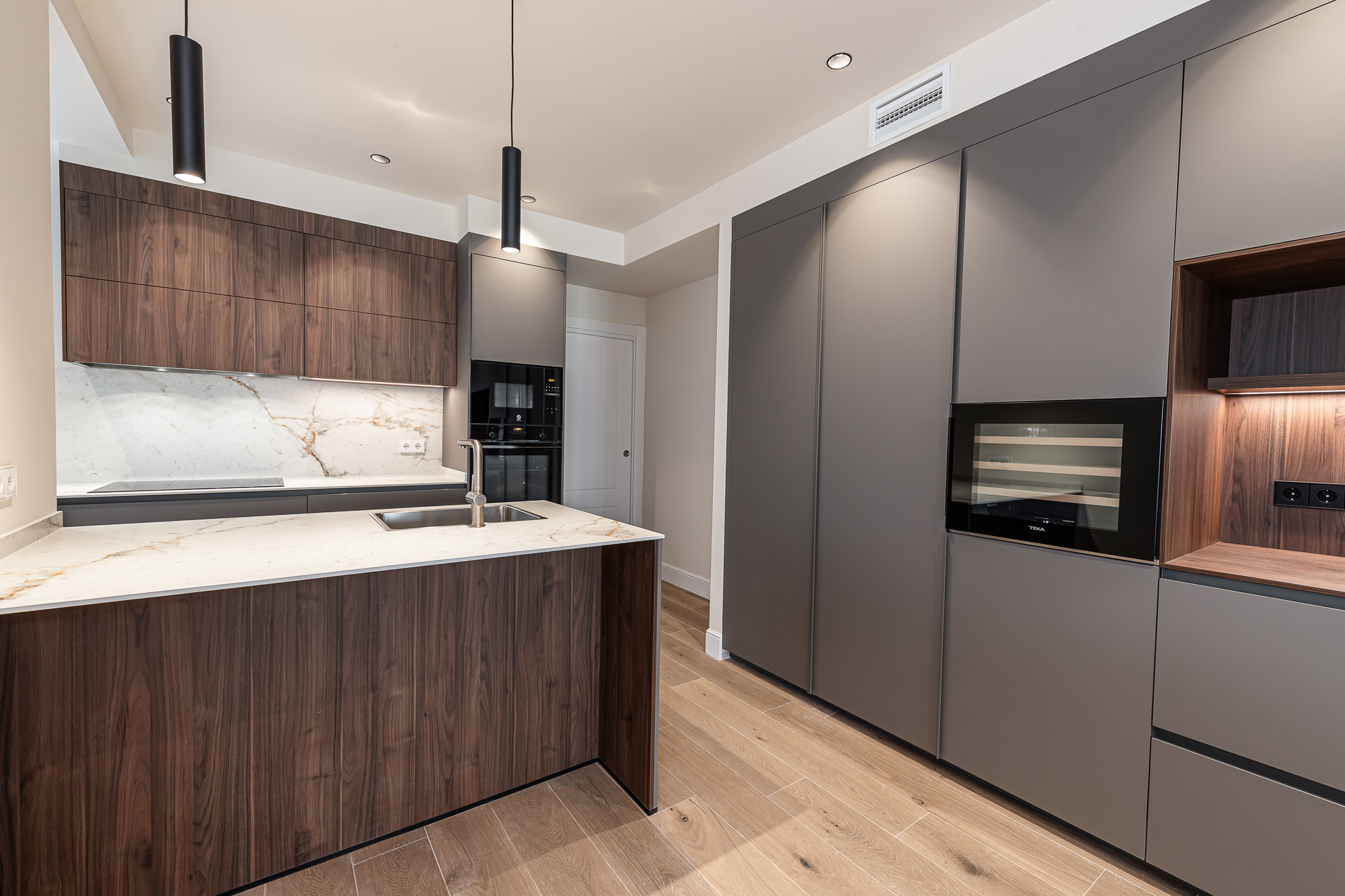Cocina con isla de madera oscura, frente veteado claro y armarios altos grises con electrodomésticos integrados