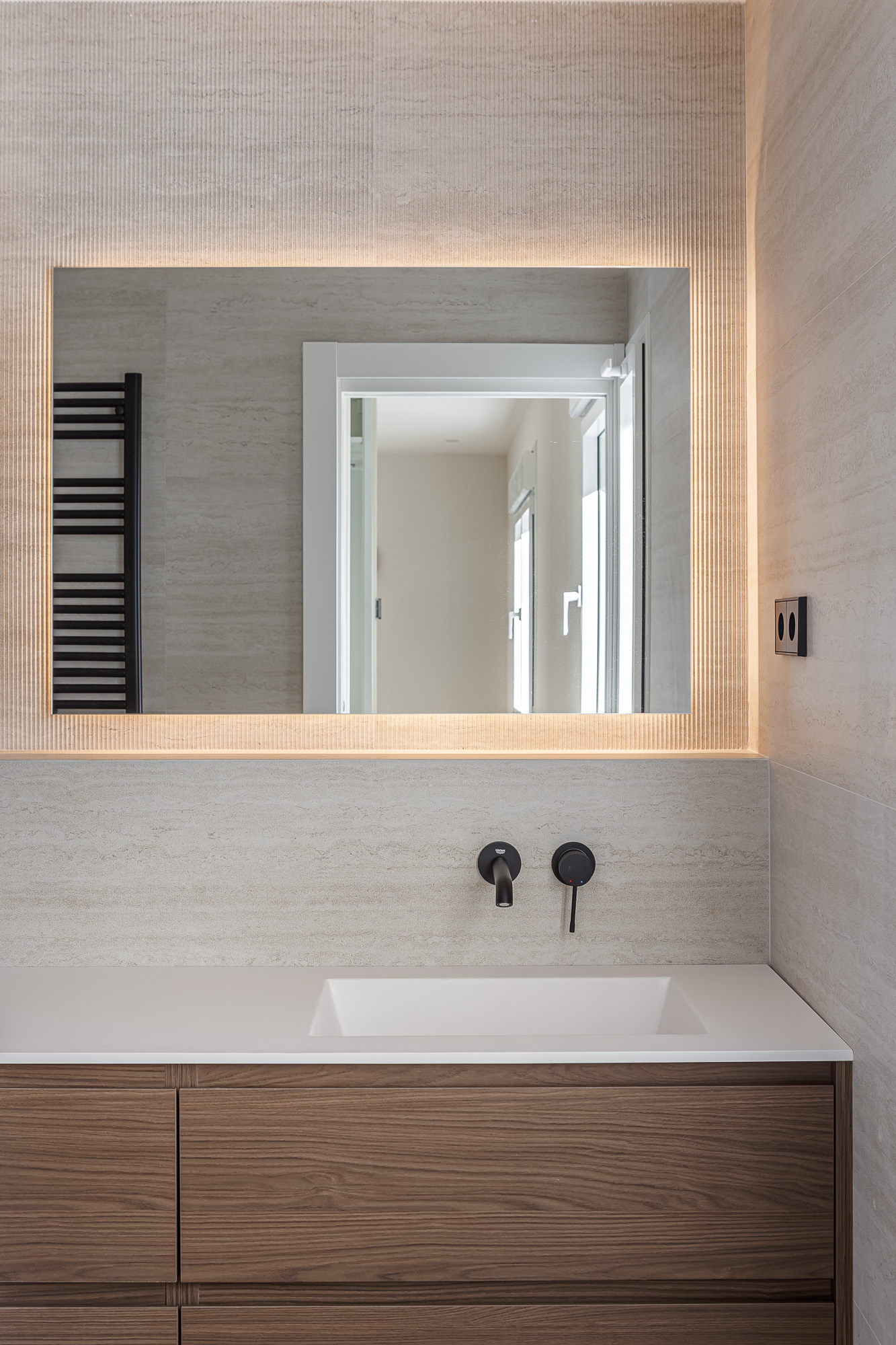 Mueble de baño de madera con encimera blanca, grifería negra de pared y espejo retroiluminado