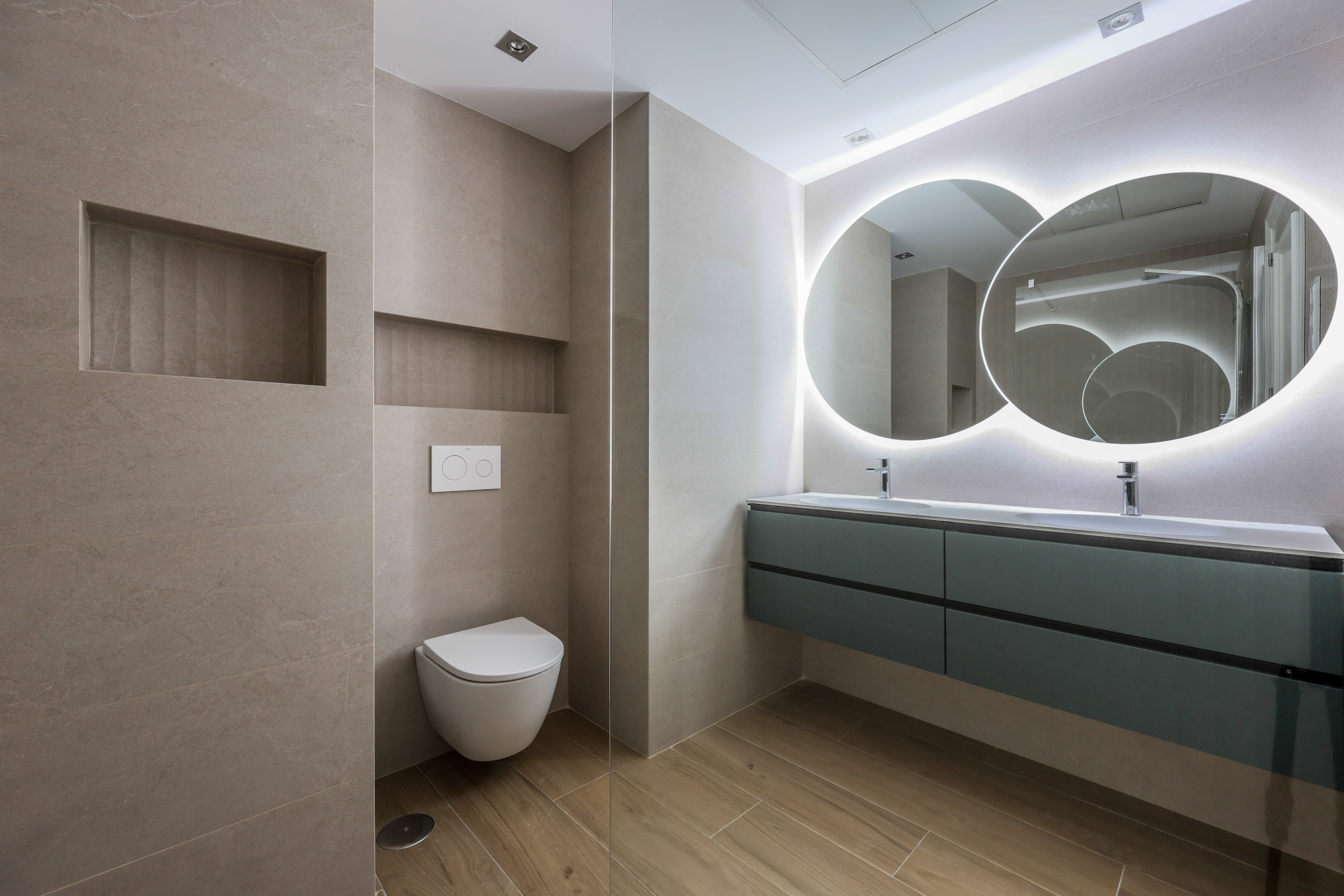 Baño beige con doble lavabo, mueble verde y espejos redondos retroiluminados