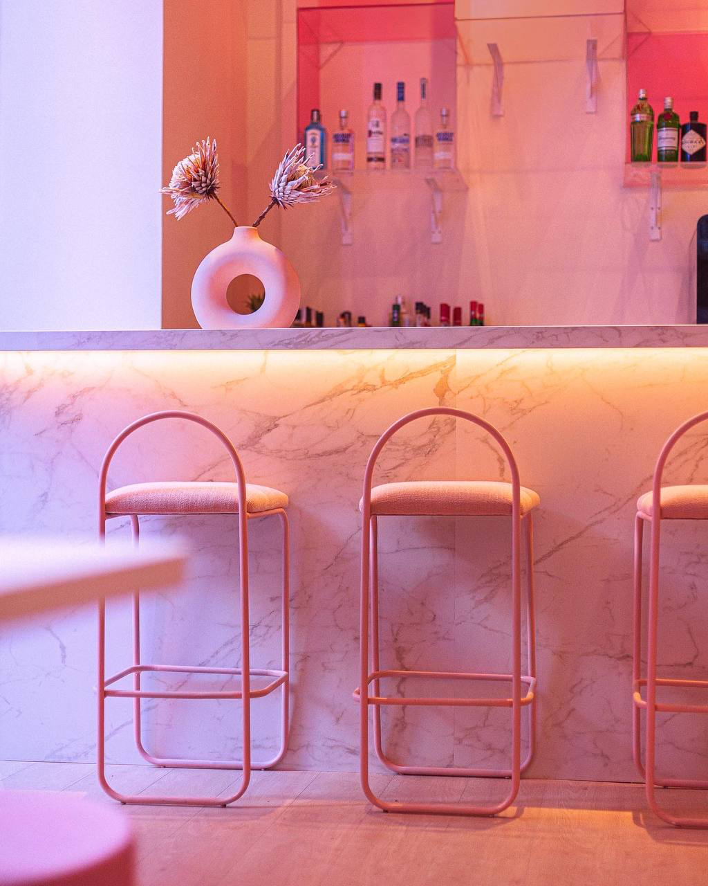 Barra con frente veteado en blanco, taburetes rosas y estantes de botellas con luz indirecta