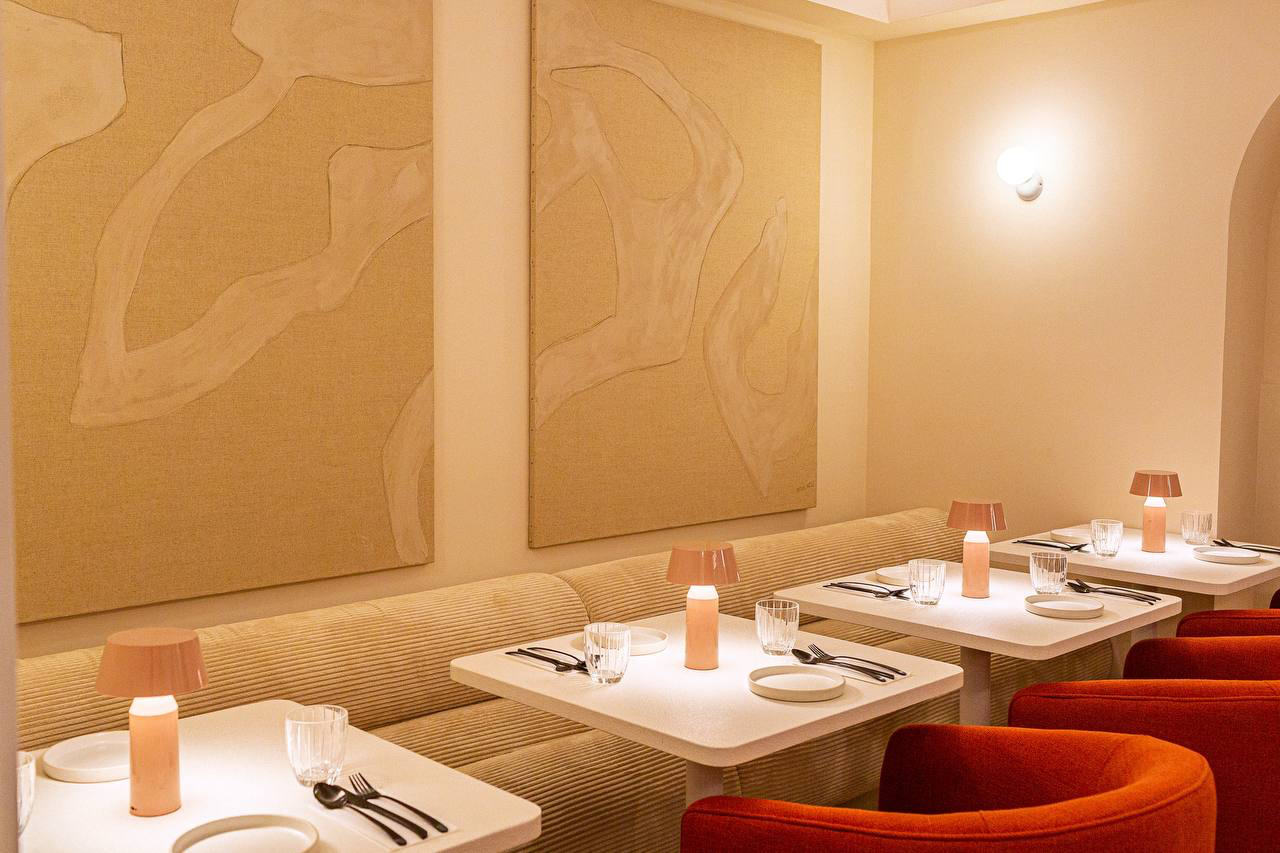 Banqueta de pana beige bajo dos paneles de lienzo con relieve, mesas blancas y sillones rojos