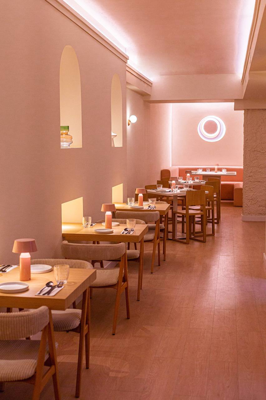 Sala alargada con hornacinas en arco, mesas de madera y banqueta naranja al fondo bajo un aplique circular
