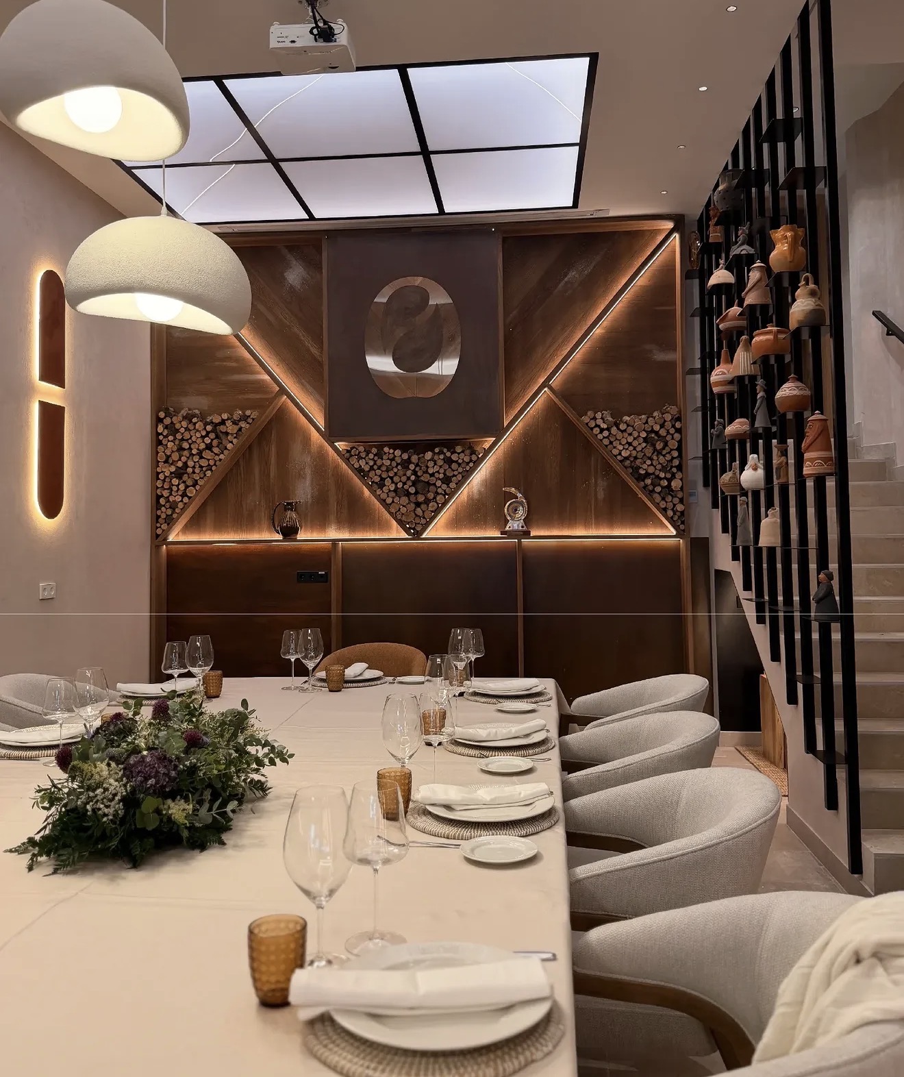 Sala con mesa larga de madera oscura puesta, sillones tapizados en tejido claro, lámparas colgantes blancas y pared de madera con iluminación lineal — Restaurante Olar by O'Grelo