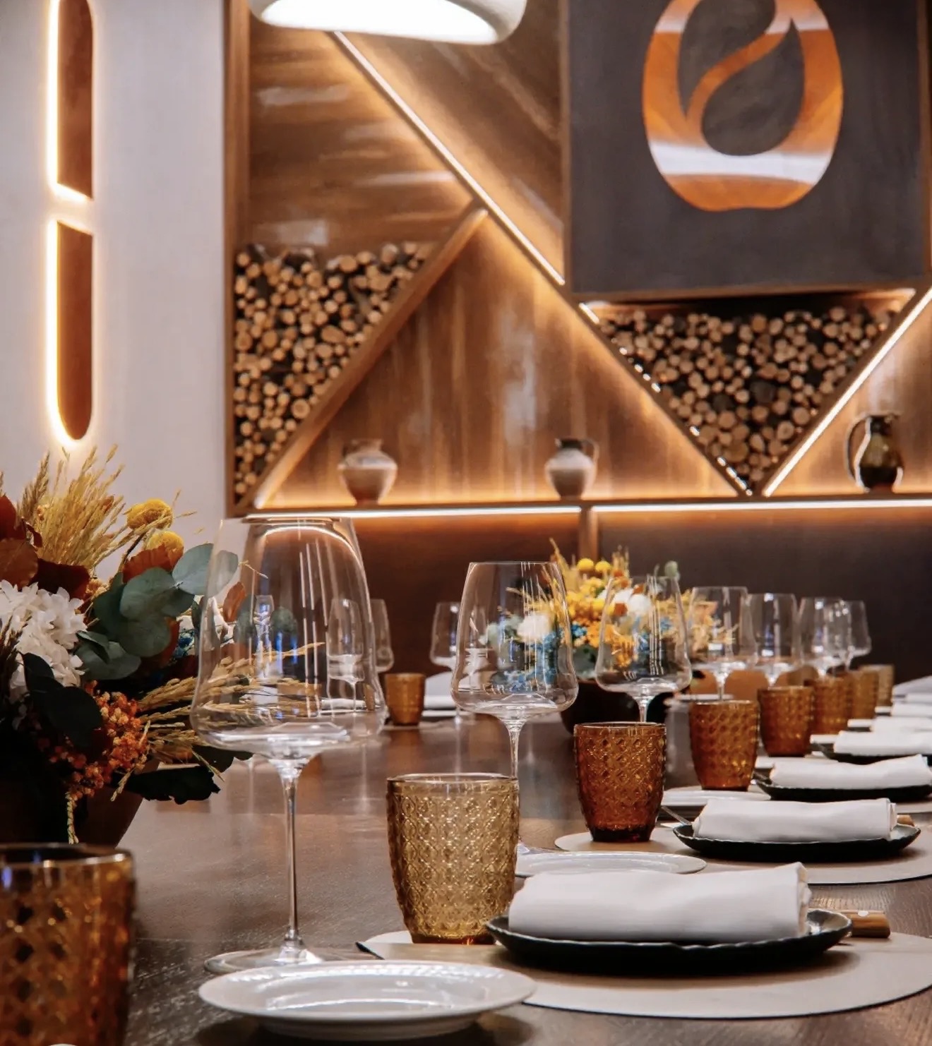Sala con mesa larga puesta, pared de madera con nichos de leña apilada y, a la derecha, escalera con piezas de cerámica sobre estructura metálica negra