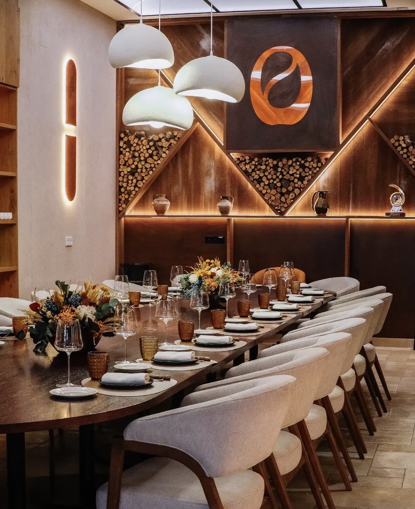 Vista a lo largo de la mesa con lámparas colgantes blancas, sillones tapizados y pared de madera con iluminación indirecta