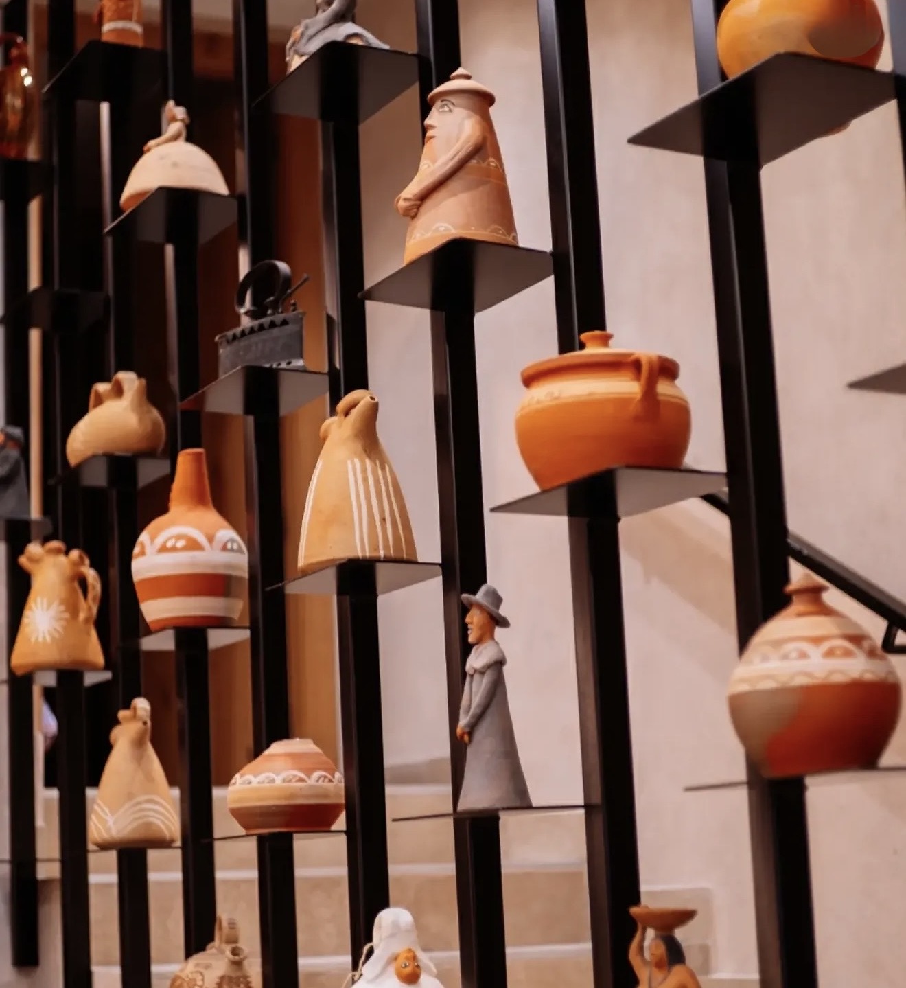 Detalle de piezas de cerámica y figuras de barro sobre baldas de metal negro junto a la escalera