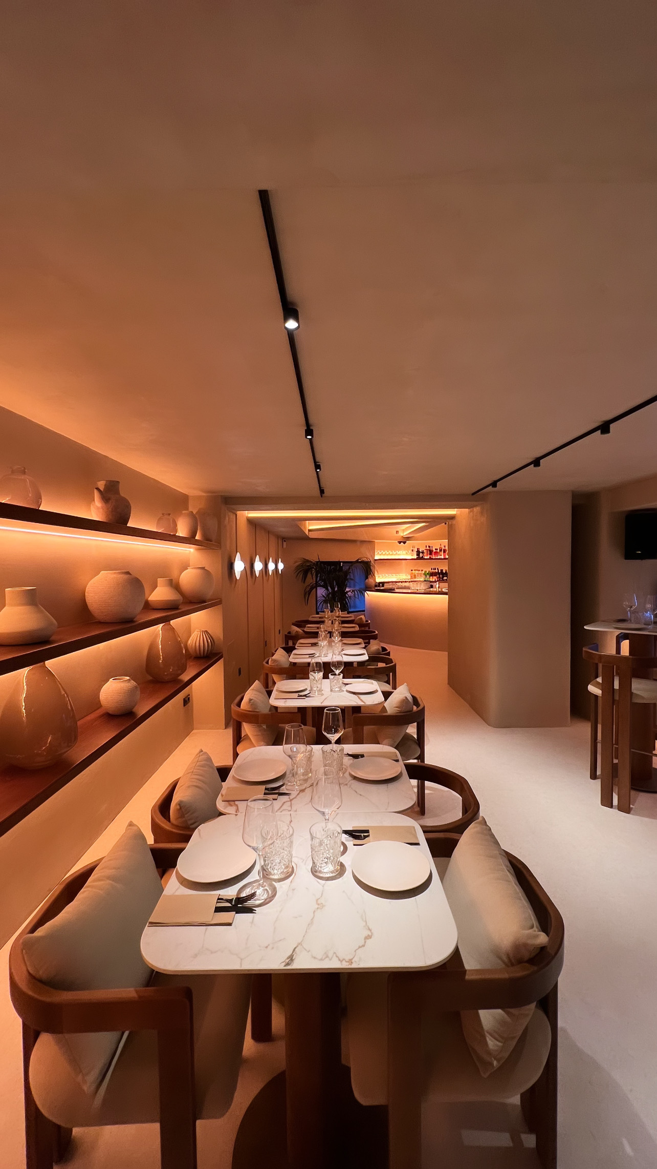 Fila de mesas de tapa blanca veteada y sillones de madera, con estantería de cerámicas iluminada a la izquierda y la barra al fondo — Restaurante Santiago Ultramarinos