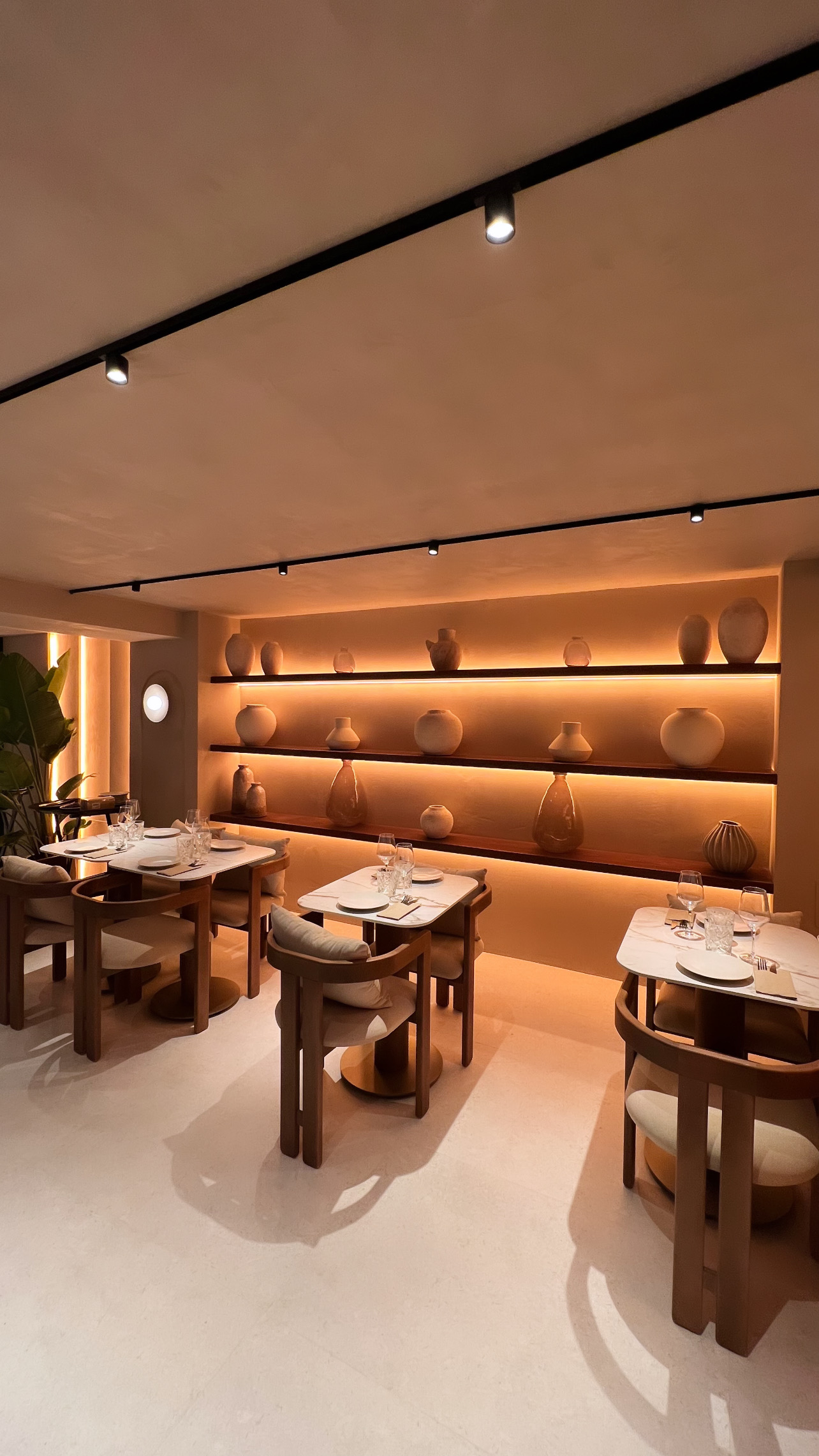 Estantería de madera con jarrones de cerámica y luz indirecta, junto a mesas de tapa veteada